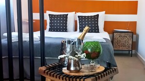Una camera d'albergo con un letto, un comodino, un tavolo con una bottiglia e dei bicchieri, e un vaso decorativo.