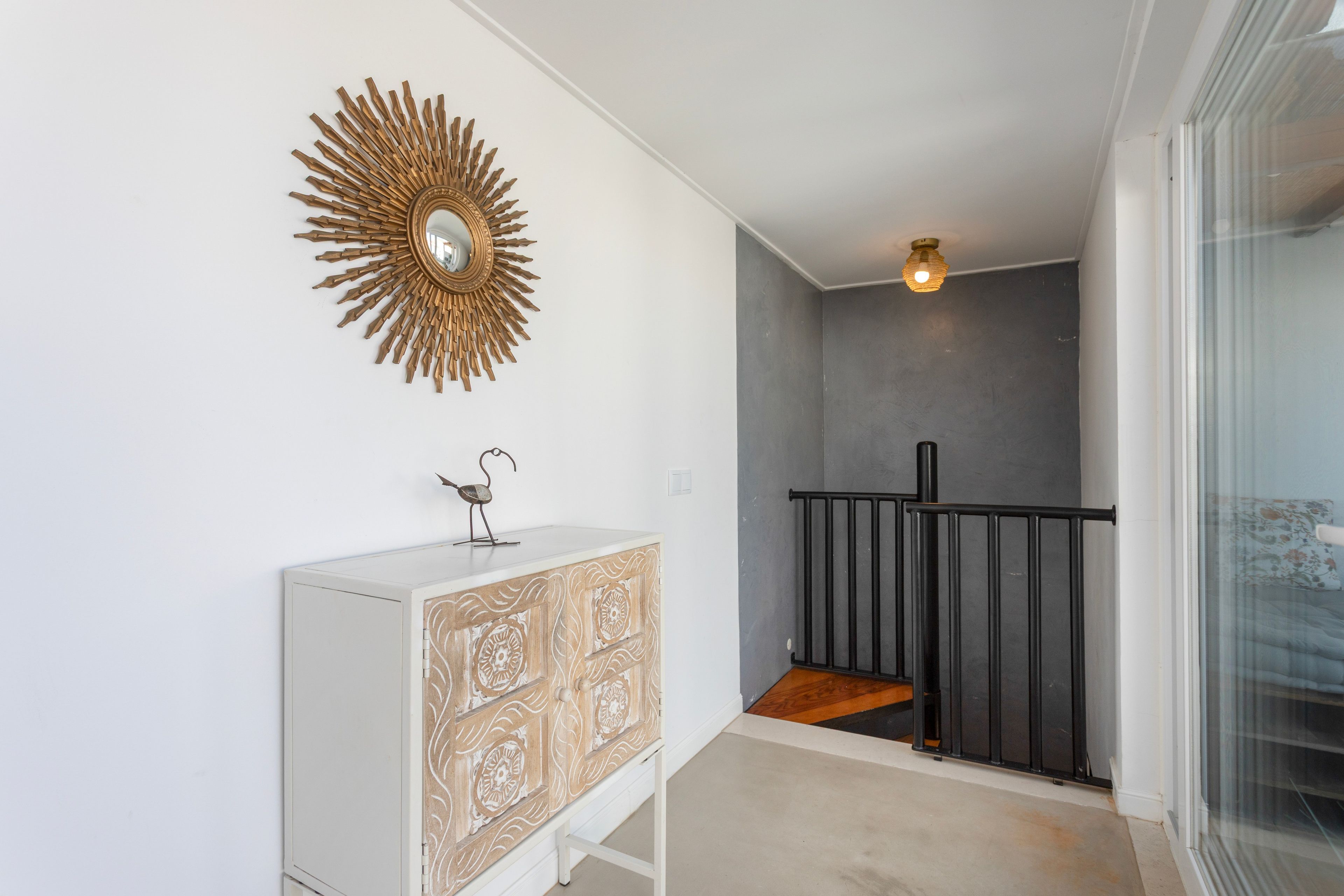 A hallway with a white cabinet, a decorative sunburst mirror, a small metal sculpture, and a staircase with a black railing.