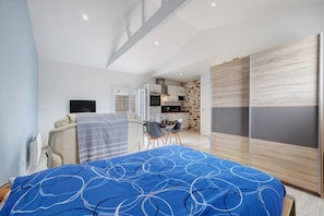 Un dormitorio con cama, escritorio y una kitchenette al fondo.