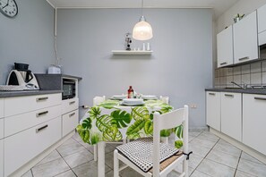 A modern kitchen with white cabinets, a dining table with a tropical leaf tablecloth, a hanging pendant light, and a clock on the wall.