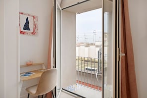 Ein Balkon mit Blick auf Häuser und eine Straße, ein Stuhl, ein kleiner Tisch mit einer Schale und ein gerahmtes Bild an der Wand.