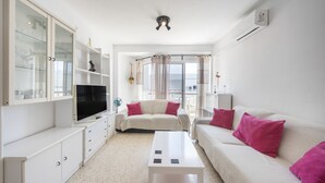 Un salon moderne avec un téléviseur à écran plat, des canapés blancs ornés de coussins roses, une table basse et un meuble vitré.