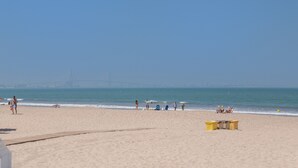 Une plage avec un ciel dégagé, un pont visible au loin, et quelques personnes ainsi que des chaises de plage.