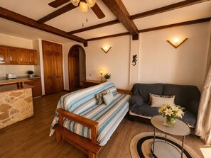Een gezellige kamer met een bed, een bank, een tafeltje met een bloevas, houten balken aan het plafond en een keuken in de verte.