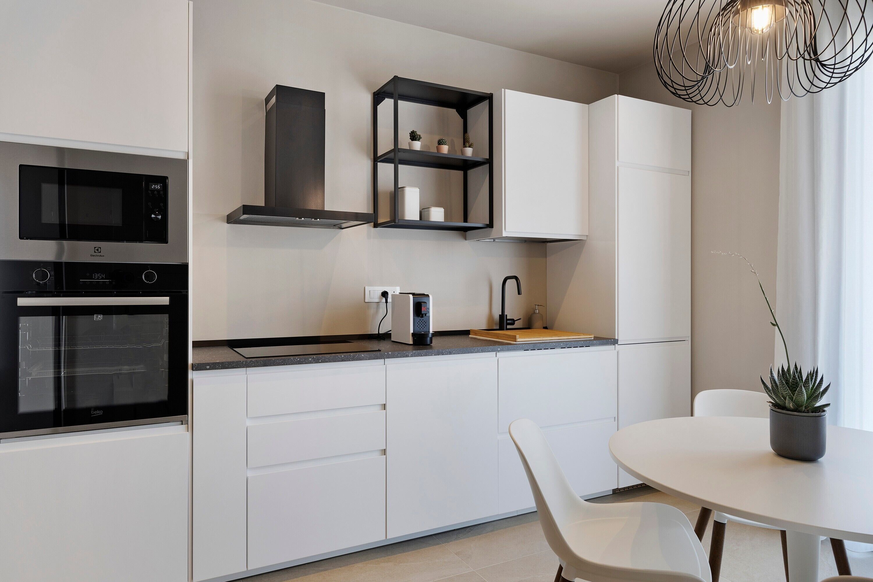 A modern kitchen with white cabinetry, a built-in microwave, a black range hood, and a small shelf with decorative items.