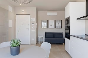 A modern kitchen with white cabinets, a grey sofa, and a dining table with a potted plant. - Apartment 'Libeccio' with Balcony, Wi-Fi and Air Conditioning (Santa Flavia)