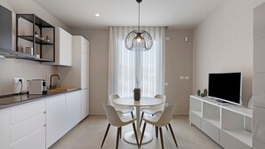 Et moderne køkken med hvide skabe, et spisebord med stole, en hængende lampe og et fjernsyn på et hvidt skab.