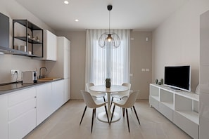 A modern kitchen with white cabinets, a dining table with chairs, a pendant light, and a TV on a white stand. - Apartment 'Libeccio' with Balcony, Wi-Fi and Air Conditioning (Santa Flavia)