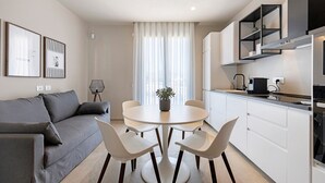 Une cuisine moderne avec des meubles blancs, un coin repas doté d’une table ronde et de chaises, et un canapé gris agrémenté de coussins.