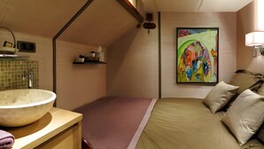 Un dormitorio compacto con un lavamanos grande tipo cuenco, una cama con muchos almohadones, un cuadro abstracto colorido y un estante pequeño con artículos de tocador.