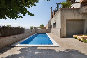 A rectangular swimming pool with a tiled deck, a closed garage door, and a stack of lounge chairs.