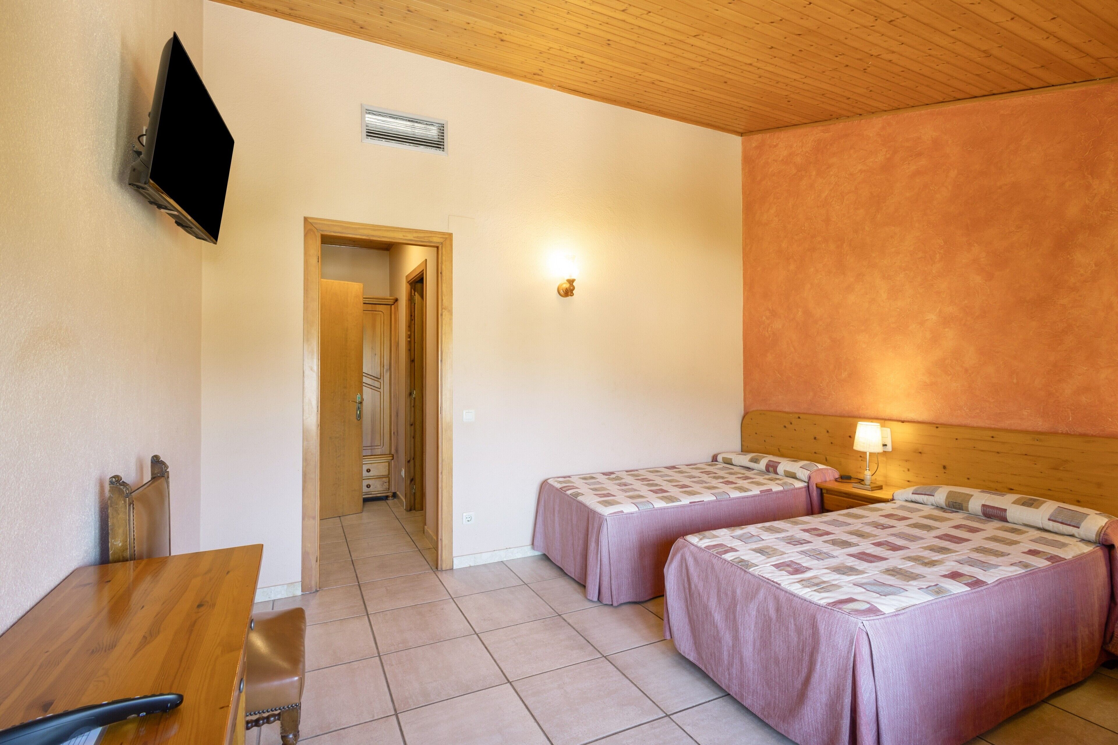A hotel room with two beds, a wooden desk, a television, and a hallway leading to another room.