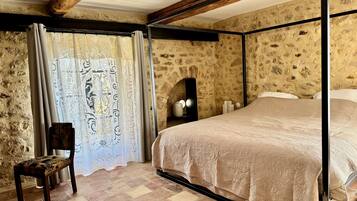 Un dormitorio con pared de piedra, una cama con colcha color beige, una silla de madera y una ventana con cortinas estampadas.