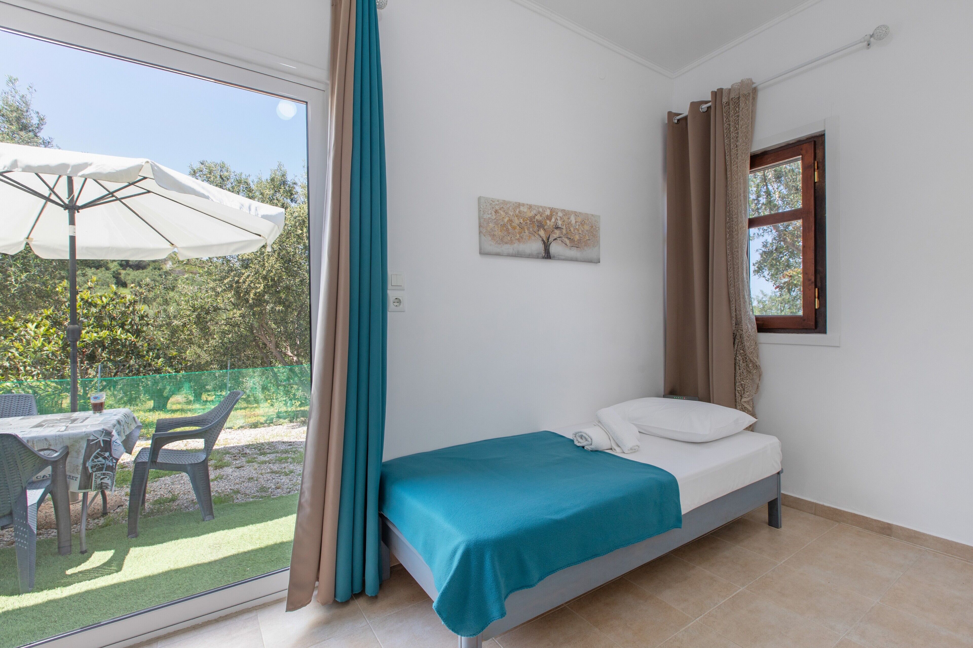 Une chambre à coucher avec un lit, une fenêtre, un tableau et une vue sur une terrasse extérieure avec une table et des chaises.