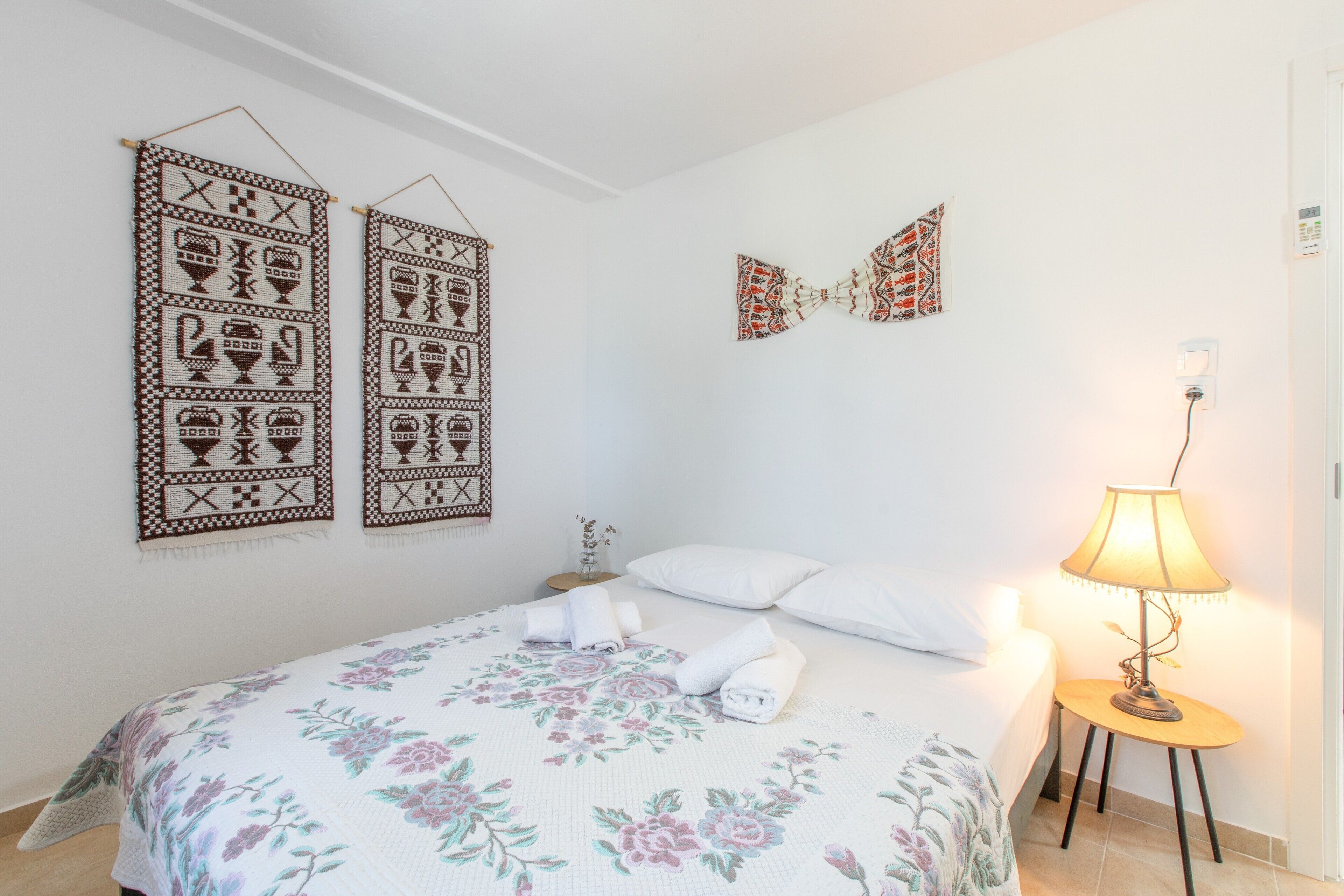 Ein ordentlich bezogenes Bett mit floralem Überwurf, zwei an der Wand befestigte Dekorationen, eine Nachttischlampe und ein kleiner Tisch mit einer Vase.