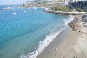 Zona costera con playa arenosa, rompeolas rocosos y varios veleros en el agua.