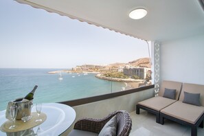 Een balkon met uitzicht op zee en een stadsbeeld, een lounge stoel, een klein tafeltje met champagne en glazen, en een decoratief wandpaneel.
