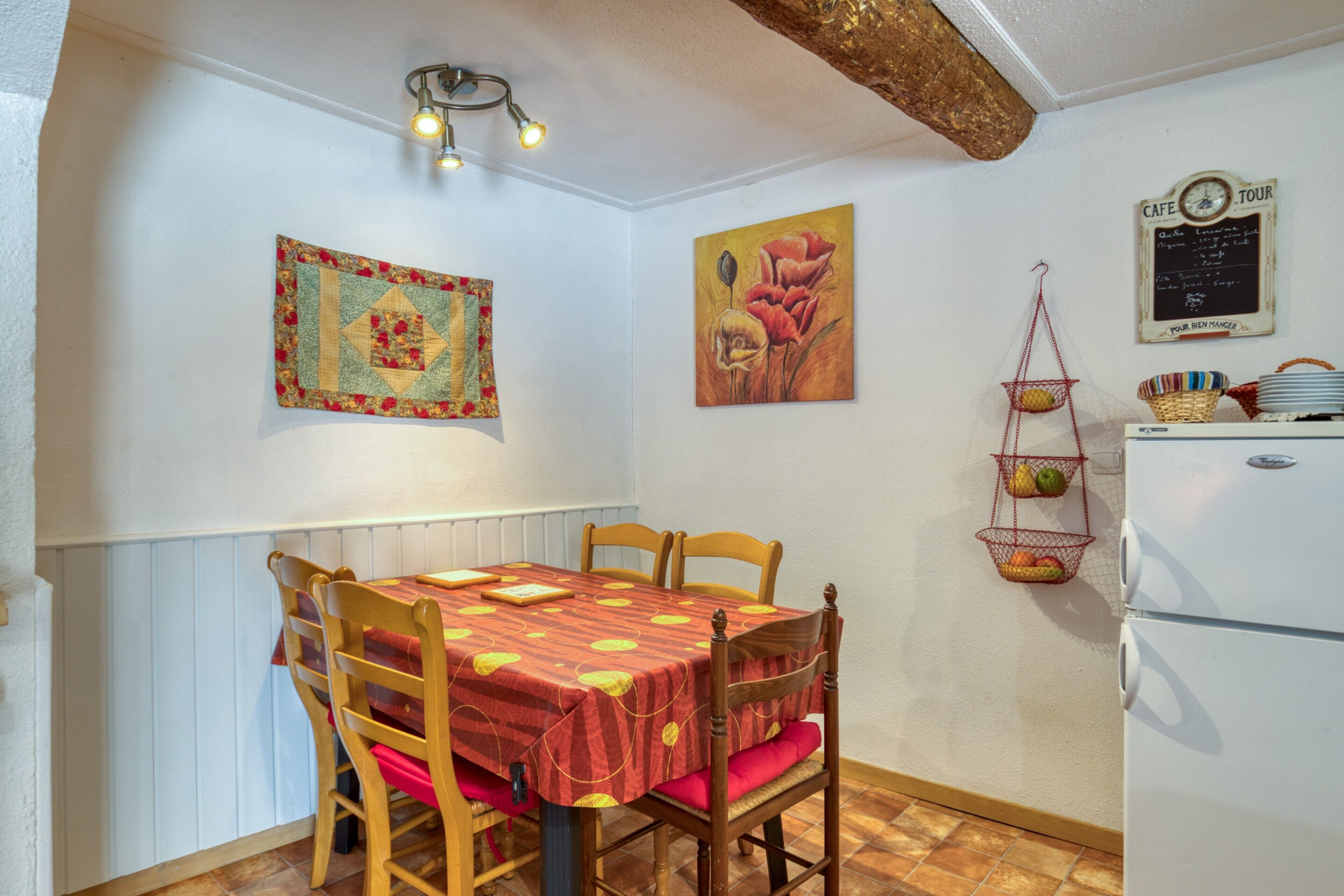 A dining area with a table set for four, a refrigerator, a wall-mounted basket with fruits, and a painting of flowers.