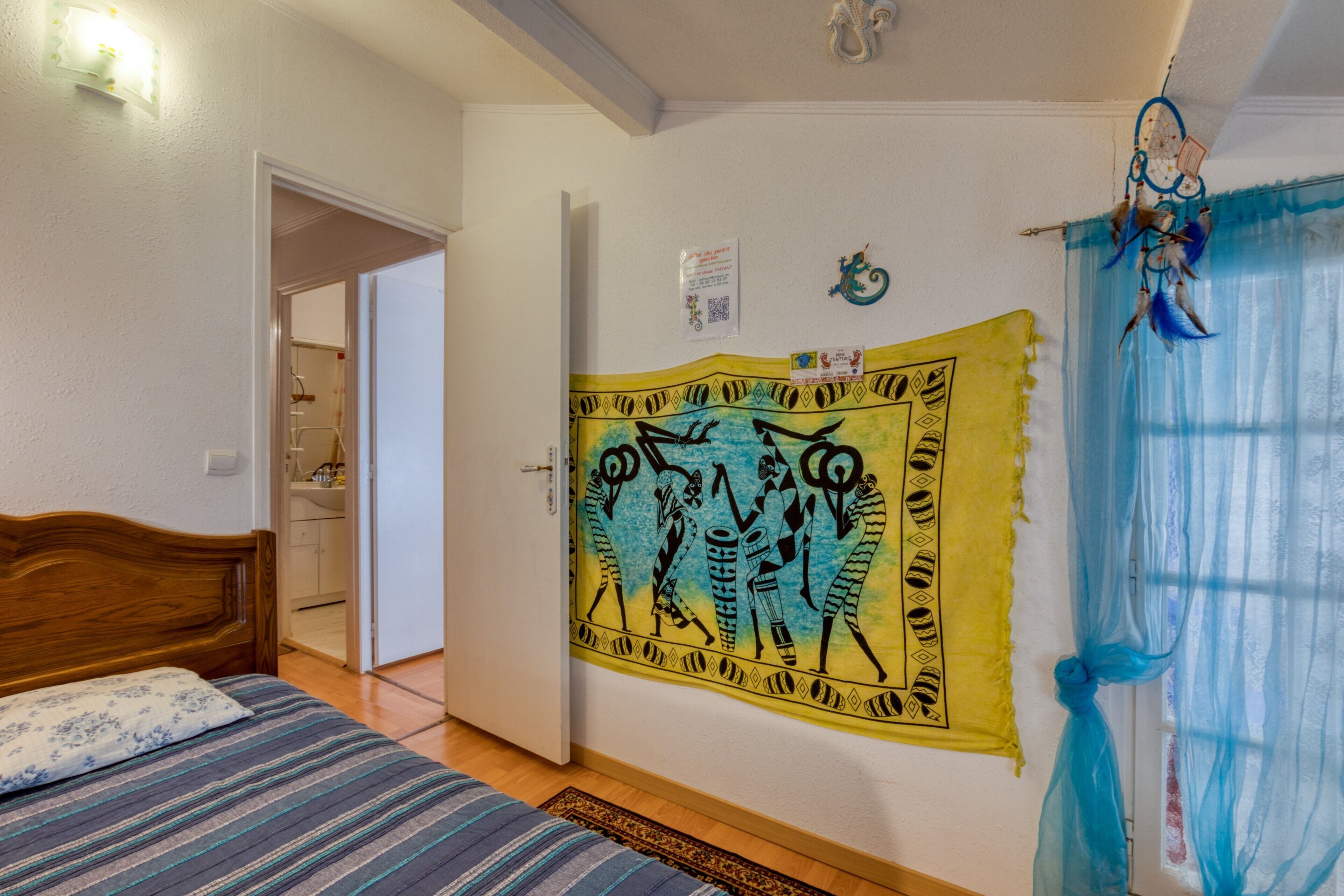Un dormitorio con una cama, un tapiz en la pared, un atrapasueños y la puerta del baño.
