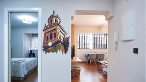 Ein Zimmer mit einem Bett, einem Schreibtisch und einem Stuhl. Es gibt ein Fenster mit Jalousien, ein an der Wand befestigtes Telefon und eine Tür zu einem weiteren Raum.