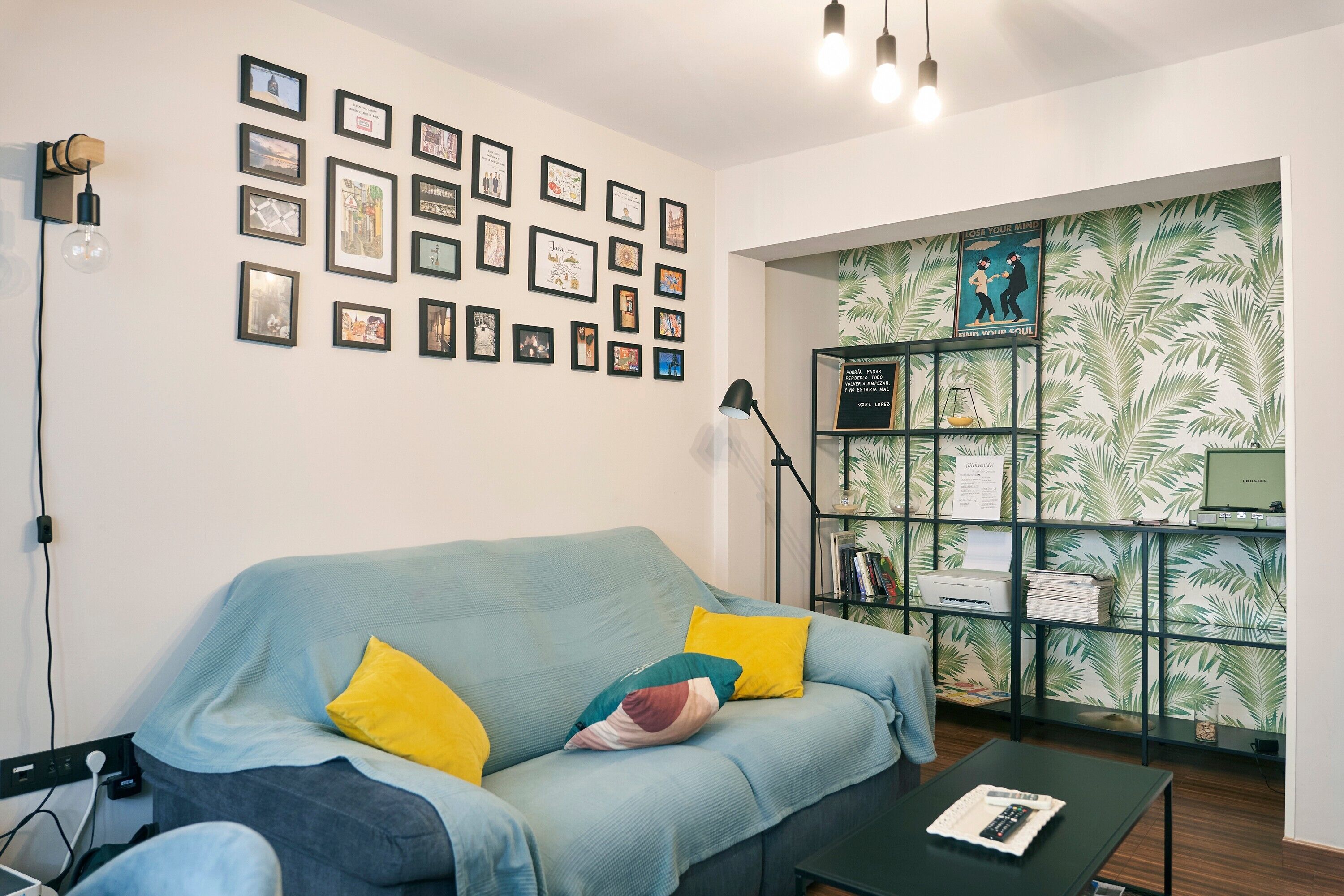 A room with a light blue sofa, yellow pillows, a black coffee table, a bookshelf, and a wall with framed pictures.