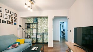 Ein Wohnzimmer mit einer Couch, einem Couchtisch und einem Fernseher, einem Bücherregal und einer Küche im Hintergrund.