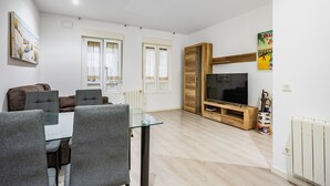 A modern living room with a glass dining table, grey upholstered chairs, a flat-screen TV, wooden entertainment unit, and a radiator.