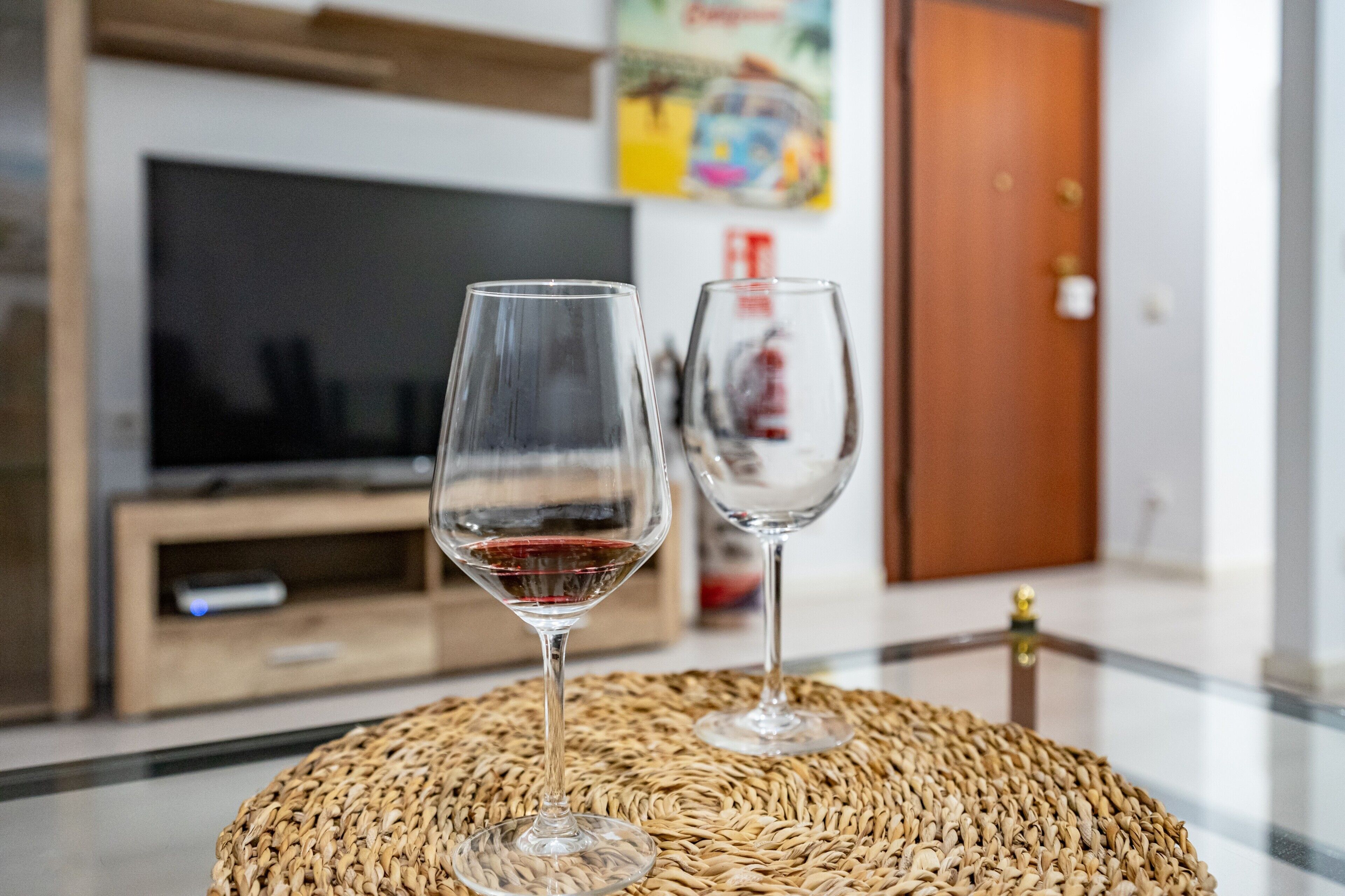Dos copas de vino vacías sobre un mantel tejido en una habitación con una televisión y una puerta de madera al fondo.