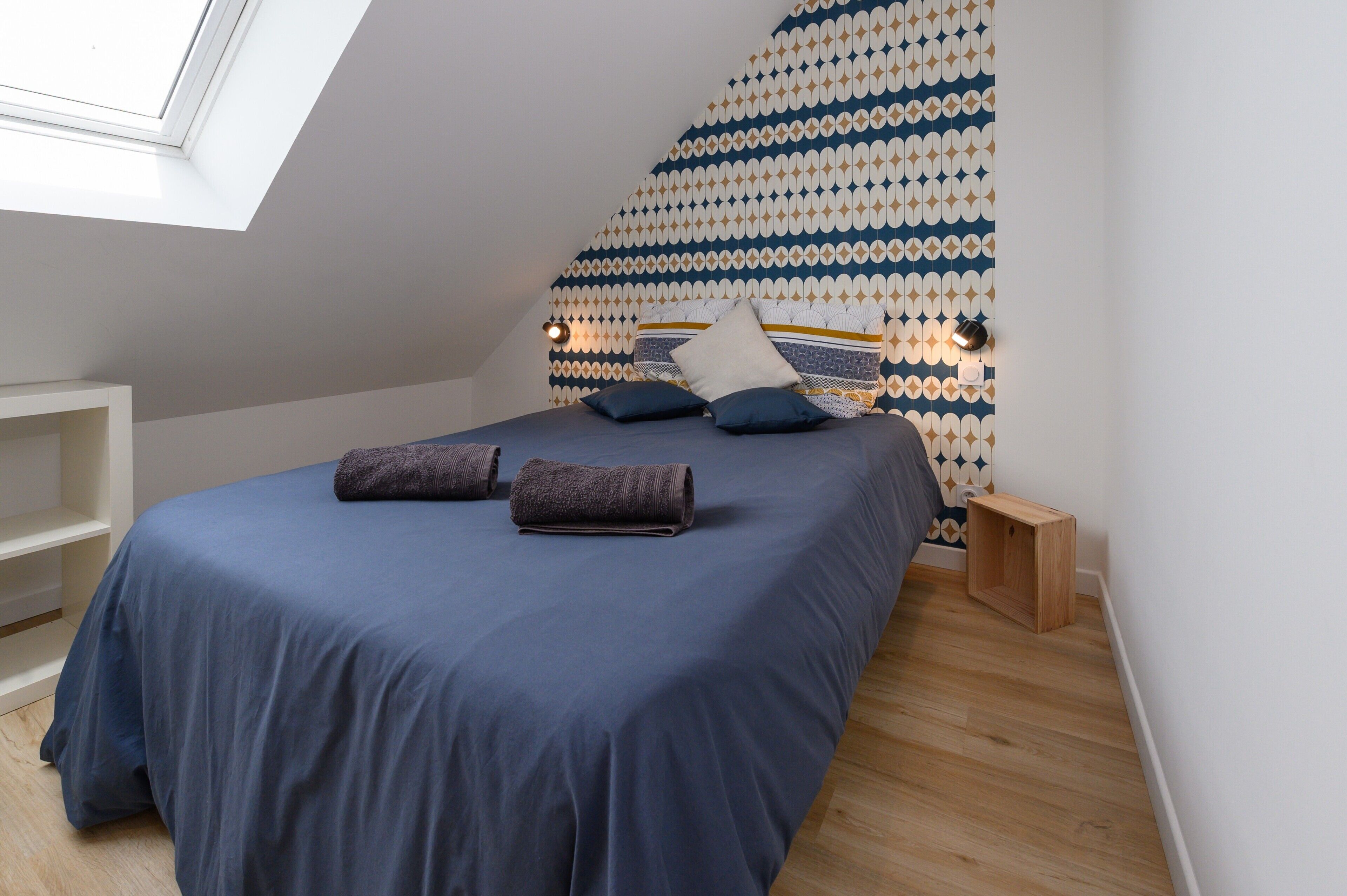 Een slaapkamer met dakraam, een behang met patroon, een bed met blauw beddengoed en een klein houten nachtkastje.