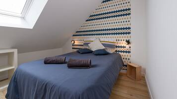 Een slaapkamer met dakraam, een behang met patroon, een bed met blauw beddengoed en een klein houten nachtkastje.
