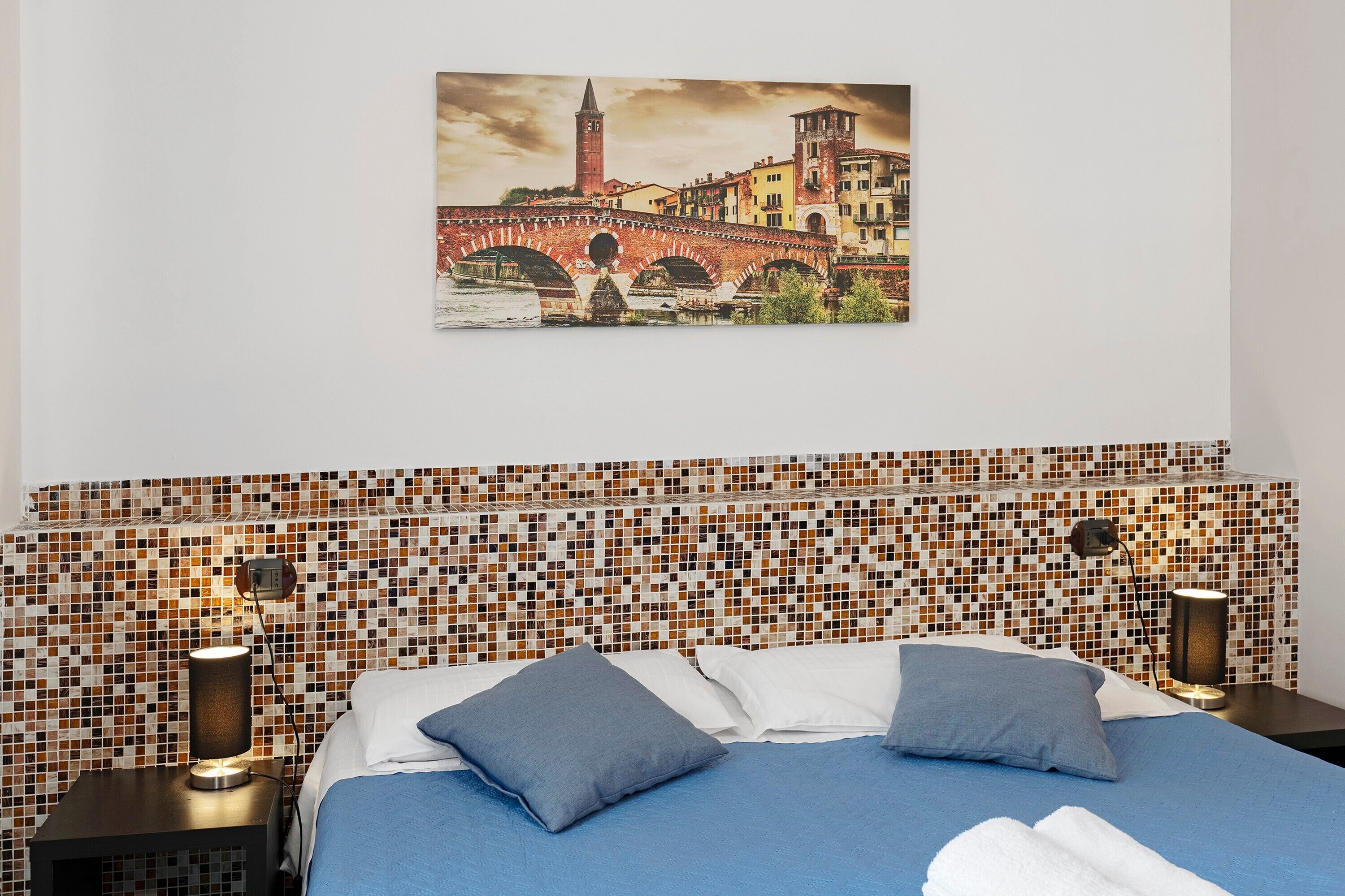 A hotel room with a bed, two bedside tables, a wall-mounted picture of a bridge, and a mosaic-patterned headboard.