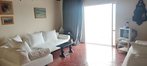 Ein Wohnzimmer mit einem weißen Sofa, einem kleinen Tisch, einem Fernseher und einem Fenster mit Vorhängen.