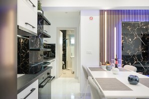 A modern kitchen with black marble countertops, a white dining area with a table, and a view of the bathroom. - Sunset Legacy B Apartment with Balcony, Wi-Fi and Air Conditioning (Rhodes)