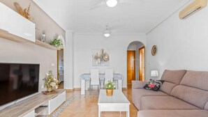 Uma sala de estar moderna com uma TV de tela plana, um sofá, uma mesa de centro e um espaço de jantar com cadeiras e mesa.