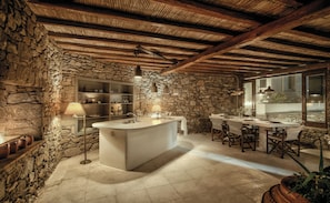 Ein rustikales Interieur mit einer Steinwand, einer zentralen Kochinsel mit Spüle sowie einem Essbereich mit Stühlen und Tischen.
