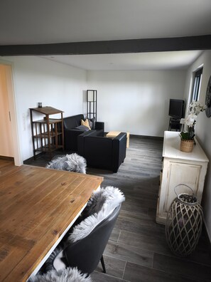 A modern living room with a wooden dining table, black sofas, a small TV, and a decorative plant. - Holiday Home 'Haus Am Deich' with Terrace, Private Garden & Wi-Fi (Norden)