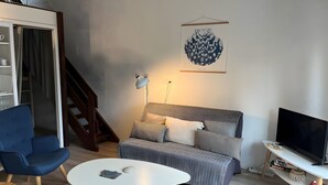 A modern living room with a sofa, a blue armchair, a white coffee table, a TV, and a staircase. - Apartment 'Cap Ferret with Pool Ref. 89' with Shared Pool and Balcony (Lège-Cap-Ferret)