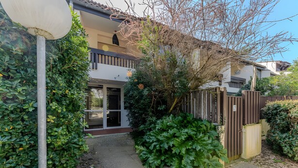 A residential building with a gated entrance, a pathway leading to the door, and dense foliage around. - Apartment 'Cap Ferret with Pool Ref. 89' with Shared Pool and Balcony (Lège-Cap-Ferret)