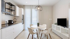Una cocina moderna con muebles blancos, una mesa de comedor con cuatro sillas, una lámpara colgante y un televisor de pantalla plana sobre un mueble blanco.