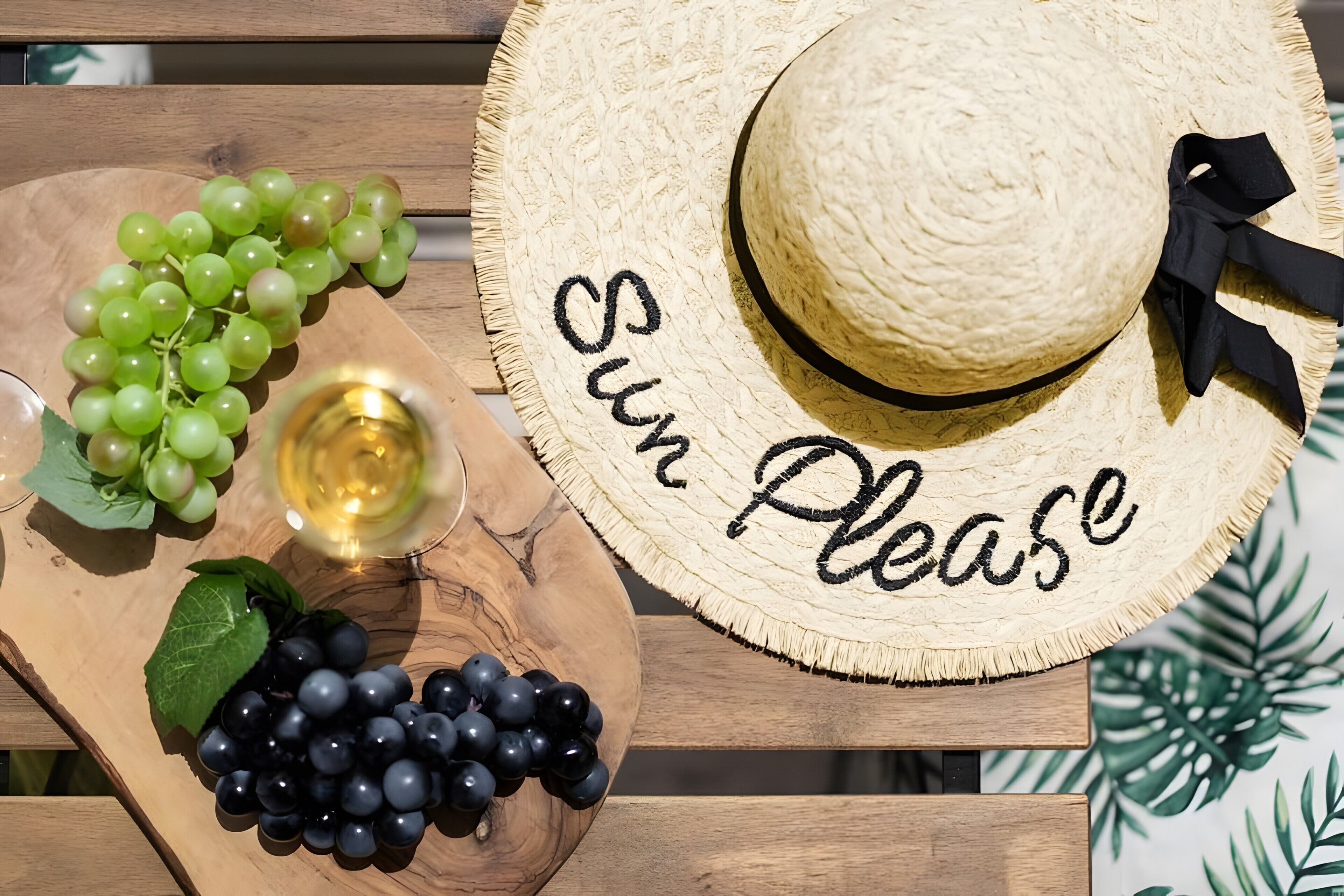 A wooden table with a straw hat, a wooden board with grapes, and a glass of white wine.