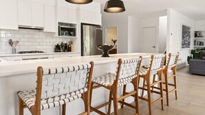 Une cuisine moderne avec des meubles blancs, un îlot central, des tabourets de bar, des luminaires suspendus et un réfrigérateur visible.