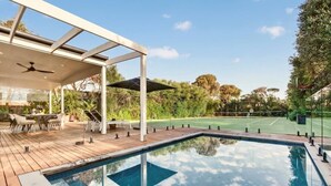 Une piscine avec une terrasse en bois, un espace salon extérieur et un court de tennis en arrière-plan.