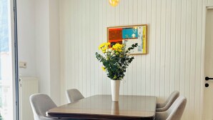Et spiseområde med et træbord, fire grå polstrede stole, en vase med gule blomster, en hængende lampe og et billede i ramme på væggen.