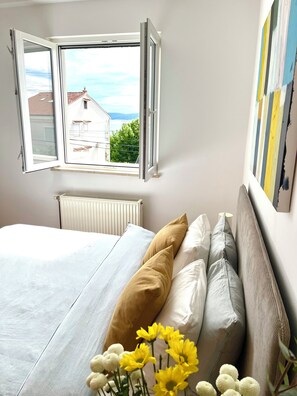 Ein Schlafzimmer mit Bett, Kissen, Heizkörper, Fenster mit freiem Ausblick und einer Wand mit einem Gemälde.