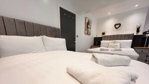 Ein Hotelzimmer mit einem großen Bett, zwei Einzelbetten, einer dunklen Tür, einem an der Wand befestigten Kunstwerk, einer herzförmigen Dekoration und einem Nachttisch mit einer Pflanze.