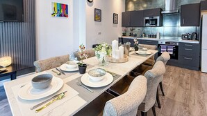 Een moderne eetruimte met een lange tafel gedekt voor vier personen, een keuken met donkere kastjes en een flatscreen televisie aan de muur.