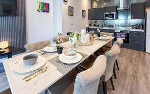 A modern dining area with a long table set for four, a kitchen with dark cabinets, and a flat-screen TV mounted on the wall. - Yellow Submarine Apartments - Sleeps 19 (Liverpool)