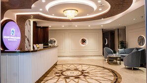 A reception area with a grand chandelier, a reception desk, a circular patterned floor, and a seating area with sofas. - Grand Ambassador Business Hotel (Kathmandu)
