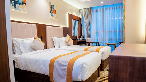 A hotel room with two beds, a desk, a chair, a coffee maker, and a large window with curtains. - Grand Ambassador Business Hotel (Kathmandu)