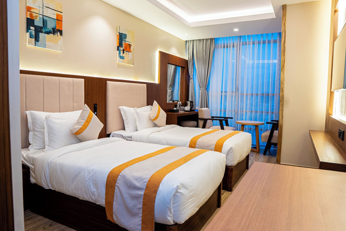 A hotel room with two beds, a desk, a chair, and a large window with curtains.